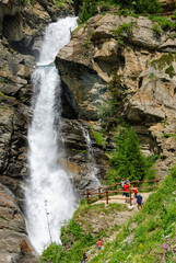 Tourists admiring the powerful Lillaz waterfalls near Cogne, Italy, with cascading alpine water, rocky cliffs and lush greenery on a bright summer day in the mountains.