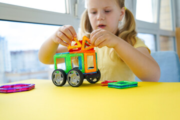 A young girl concentrates on building with magnetic construction tiles, creating imaginative designs against a solid yellow background surface.