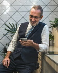 business man in formal suit and tie leaning against desk while use smartphone in modern office with geometric wall tiles and green plant. image conveys professionalism and contemporary work culture.