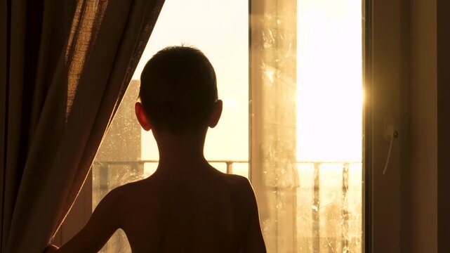 Back view of silhouetted boy opening curtains to look out window, with golden morning sunlight streaming into room. Child creating warm atmosphere of new day. Urban landscape from window at sunset