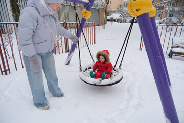 Child dressed in a red winter coat sits joyfully on a swing, surrounded by snow in a park. The...