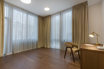Fototapeta premium Bright room has large windows covered with light curtains. A wooden desk and a chair are placed on the wooden floor. The room appears empty and open, suitable for various activities