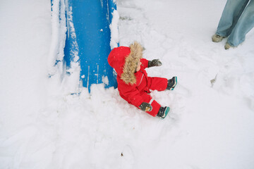 Child wearing warm red winter clothes sits on a snowy playground slide. Surrounded by snow, the child appears calm and comfortable in a winter play setting, enjoying the outdoor environment.