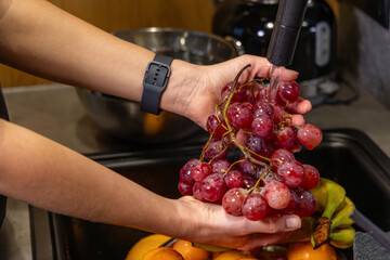 Person washing red grapes under kitchen faucet. Close-up of hands rinsing a bunch of fresh red grapes under running water in a kitchen sink, preparing healthy fruit to eat.