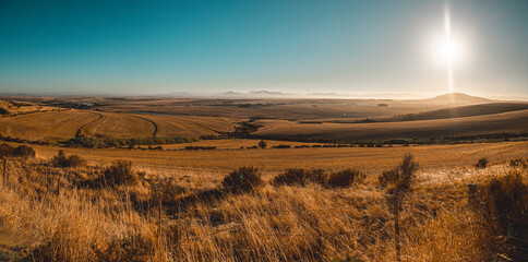 A vibrant golden morning floods a sprawling rural landscape with warm light. In the foreground, harvested fields roll toward a distant, hazy city skyline nestled under a bright, glowing sun.
