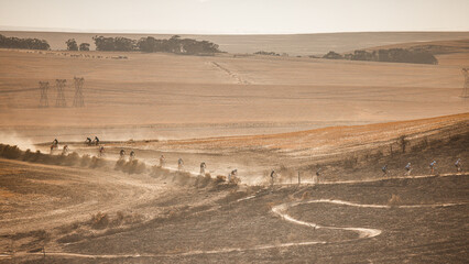 A long line of mountain bikers snakes up a dusty, winding trail during the race. Set against a hazy backdrop of layered mountain ridges and rolling hills, the scene highlights the grueling endurance