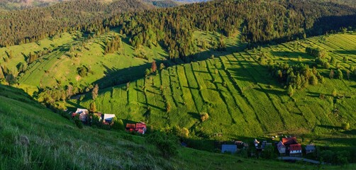 Scenic countryside landscape in Slovakia with green hillside meadows, patchwork farmland and a small rural village with traditional houses, forested slopes and warm evening light creating long shadows © Ivan