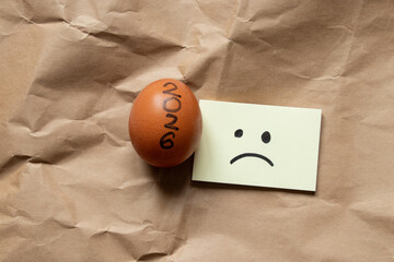 A brown chicken egg with the inscription 2026 and a sad picture drawn on yellow paper lies on crumpled craft paper, a symbol of the New Year, time, food