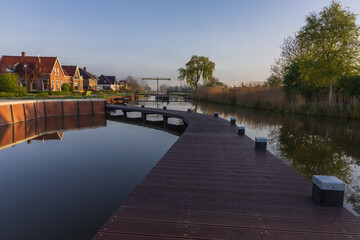 Fototapeta premium Modern wooden boardwalk along the canal in Bedum, Netherlands. Morning haze, historic brick houses and a traditional drawbridge in distance