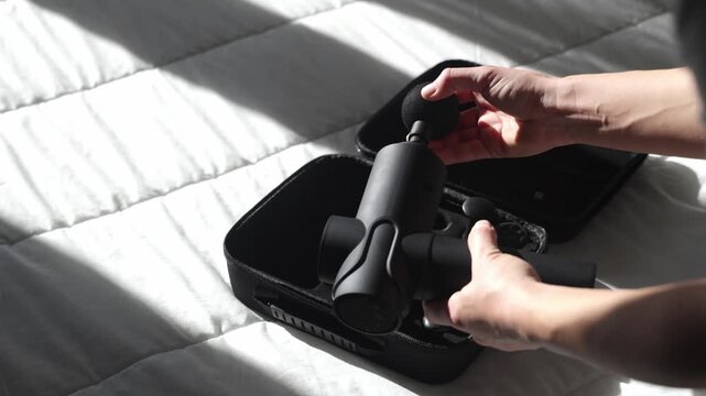 Black massage gun is being removed from a carrying case on a soft white bedspread, showcasing the unpacking process and visible attachments for muscle recovery