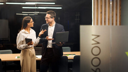 Two young colleagues having conversation using laptop and tablet behind glass wall in board room, creative director and project manager discussing work