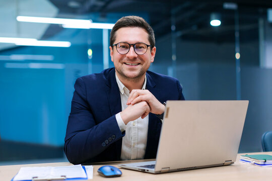 Charismatic European businessman sitting at desk in stylish office working on laptop and ready for business, smiling at camera