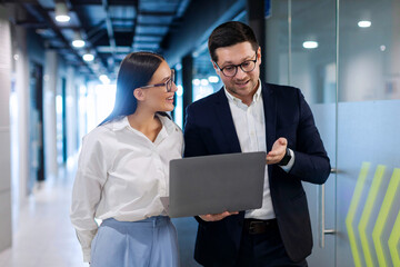 Business designers discussing project, looking at laptop monitor screen, man and woman colleagues talking during break, standing at office hallway