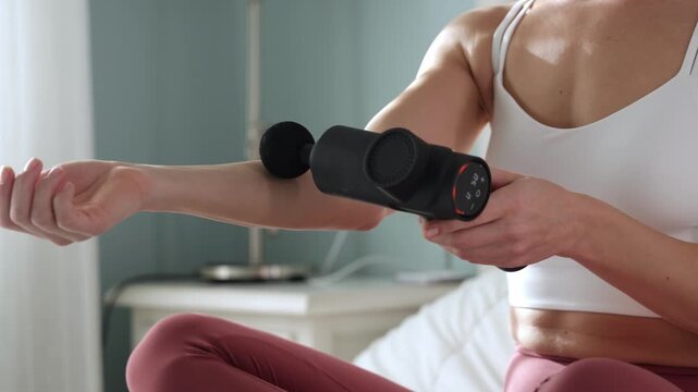 Woman using a vibrating massage gun on her arm while seated on a bed, showcasing the massage device in action with visible controls and ergonomic design