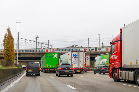 Belgium - November 28 2025: Heavy Traffic with Multiple Logistics Trucks from Heppner, HC Transports and Fleur Passing Under a Railway Bridge with a Commuter Train in Antwerp, Belgium