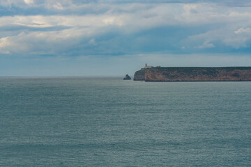 Lighthouse stands at the edge of a far-off cliff surrounded by calm ocean and clouds.