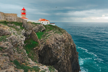 Red-roofed lighthouse stands atop rugged cliffs above the blue Atlantic waters under dramatic sky.