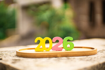 Colorful 2026 wooden numbers on a tray over a stone surface with lush green bokeh. Perfect for New Year 2026 and future planning themes