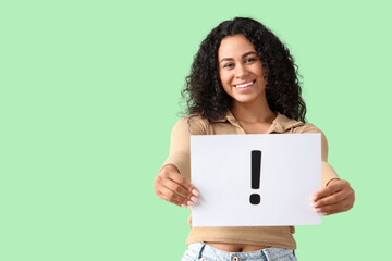 Happy young African-American woman holding paper sheet with exclamation mark on green background
