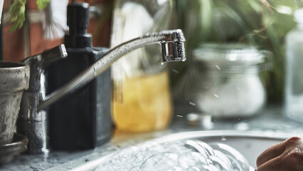 Person washing dishes with running water at kitchen sink in daytime near plants and kitchen items.