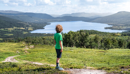 A boy with curly blond hair looks out from a hiking trail on a mountainside. He sees a lake surrounded by mountains in Trollheimen, Norway.
