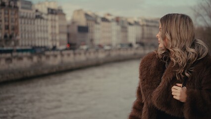 A woman wearing a brown fur coat stands overlooking the Seine River in Paris France with a cityscape background. This image is suitable for travel tourism and romantic getaway themes.