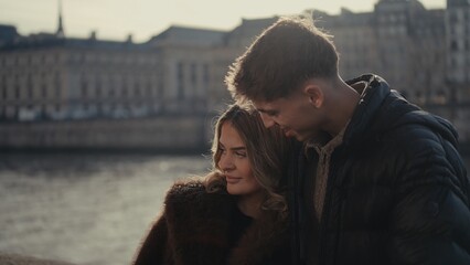 A romantic couple is embracing in Paris France with the Seine River and classic architecture in the background The young woman and man are enjoying the cityscape and their travel destination togethern