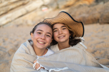 Smiling young girls wrapped in blanket sitting together on sandy beach enjoying warmth and peaceful coastal atmosphere