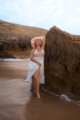 Smiling woman in summer dress and hat leaning on large rock on sandy beach near sea enjoying vacation during sunny day