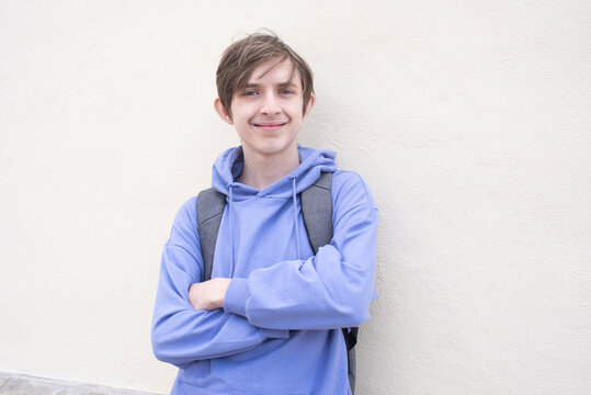 A portrait of a smiling teenager in a lilac hoodie with a backpack. The child has his hands crossed and is standing against a white wall.