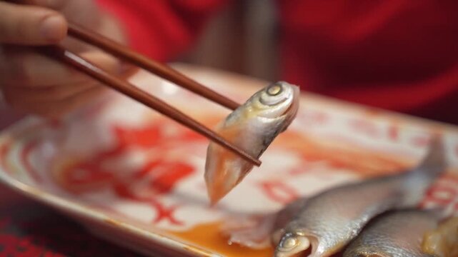 The Last Bite of Fish. Chinese New Year. A pair of chopsticks gingerly picking up the very last, small piece of fish from the serving platter, symbolizing completion and wishing for surplus.
