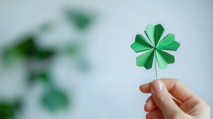 A hand holding a green four-leaf clover against a blurred background of leaves and white wall