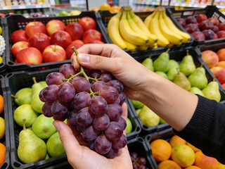 A close-up shot of hands holding a fresh bunch of red grapes surrounded by apples, bananas, and pears at a supermarket.