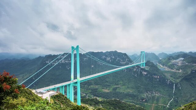 time-lapse of the Huajiang Canyon Bridge in Guizhou, China, under cloudy skies. A stunning suspension bridge spans a deep mountain canyon with lush green peaks.