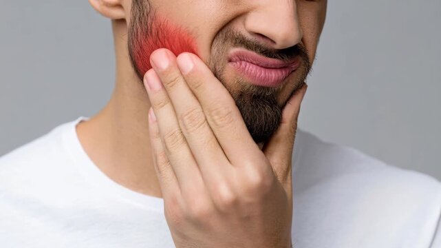 A man grimaces in pain, clutching his jaw where a red mark indicates discomfort.
