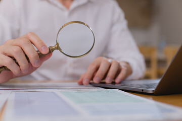 businessman using magnifying glass to examine financial documents on desk with laptop, focusing on detailed analysis, audit data verification in office setting.