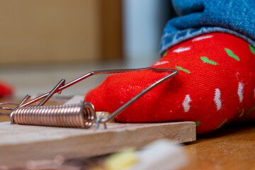 A close-up view shows a person's red festive sock-covered foot caught in a metal mousetrap on a wooden floor, with part of the person's jeans visible in the background