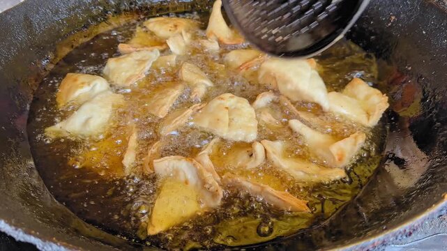 Close-up of samosas being stirred in hot oil inside a dark wok, bubbling surface and golden pastry edges shifting as metal skimmer moves through sizzling oil.