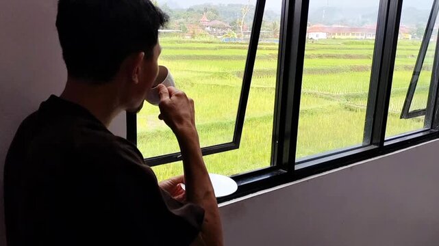 A man drinking coffee by the window for peace, quiet, or relaxation on the weekend while enjoying the view. A cup of coffee, a morning drink, or someone at home to relax with.