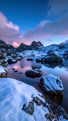 Snow-dusted rocks frame a calm lake reflecting mountains under a dusky sky with soft, pink-tinged clouds at twilight
