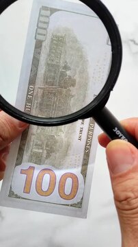 Counterfeit money. A man examines a hundred-dollar bill with a magnifying glass. Looking for watermarks on the bill. The concept of counterfeit money exchange.
