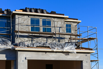 Two-story residential home under construction with exterior scaffolding and protective sheeting, showing active stucco work, window installation, and ongoing development within a growing suburban hous