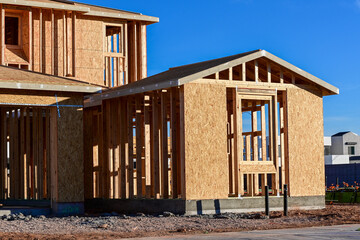 Wood-framed residential building under construction with exposed lumber and sheathing, showing early structural stages of a new home build within a growing suburban development