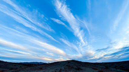 A wide open sky with streaks of white clouds stretches above a barren hilly landscape the scene is bright and calm with soft light and a sense of openness and space