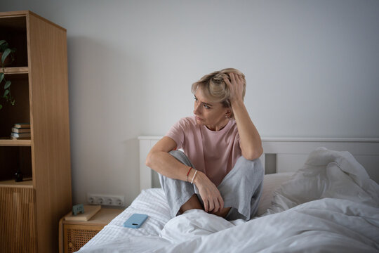 Unmotivated middle aged woman with tired expression sitting in bed in bedroom, lady woke up after sleepless night feeling fatigue, looking at window with no will to get up, seasonal apathy and boredom