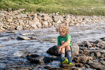 A boy sits on a rock by a river in Norway, with clear water flowing around him. He is enjoying the moment while surrounded by nature on a sunny day.
