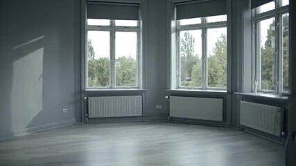 A large, empty room with gray walls, wooden flooring, and three large windows on adjacent walls allowing natural light to illuminate the space