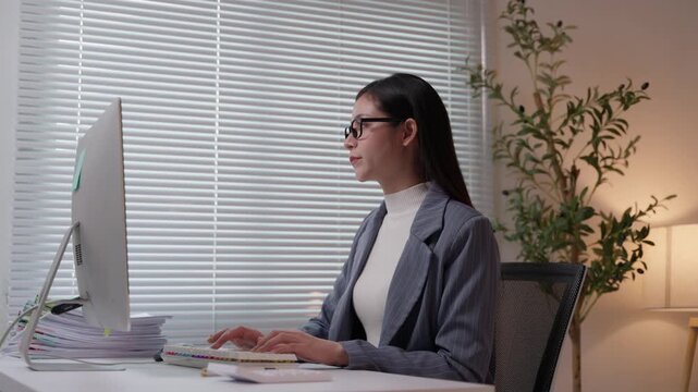 Focused asian businesswoman with glasses typing on a computer keyboard while working late in the office. Overtime and deadline concepts