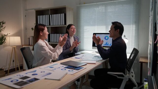 Three young Asian professionals raising hands and clapping in a modern office, celebrating a successful business deal or project completion