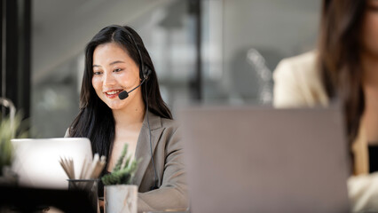 Smiling Asian woman wearing headset working in a modern office, customer service representative, call center agent or remote freelancer using laptop computer.